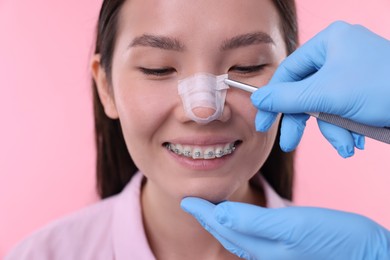 Photo of Doctor removing medical bandage from patient's nose after plastic surgery operation on pink background, closeup