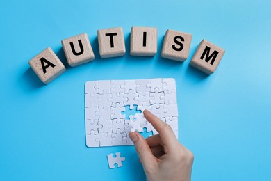 Photo of Word Autism made of wooden cubes. Woman solving puzzle on light blue background, top view