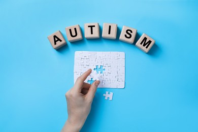 Photo of Word Autism made of wooden cubes. Woman solving puzzle on light blue background, top view