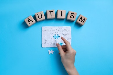 Photo of Word Autism made of wooden cubes. Woman solving puzzle on light blue background, top view