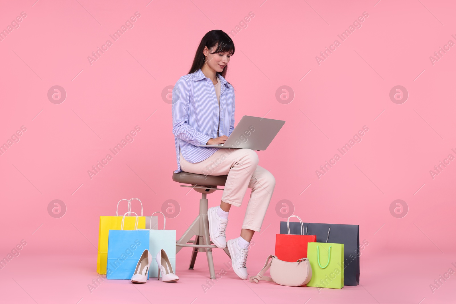 Photo of Internet shopping. Beautiful woman with laptop sitting on stool among colorful bags against pink background