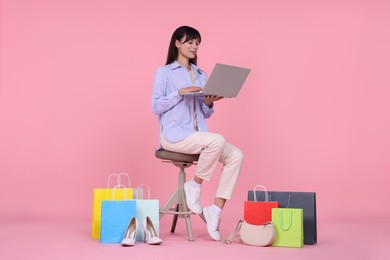 Photo of Internet shopping. Happy woman with laptop sitting on stool among colorful bags against pink background