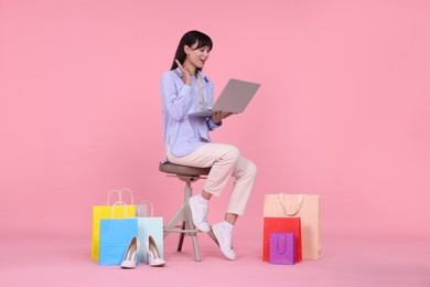 Photo of Internet shopping. Happy woman with laptop sitting on stool among colorful bags against pink background