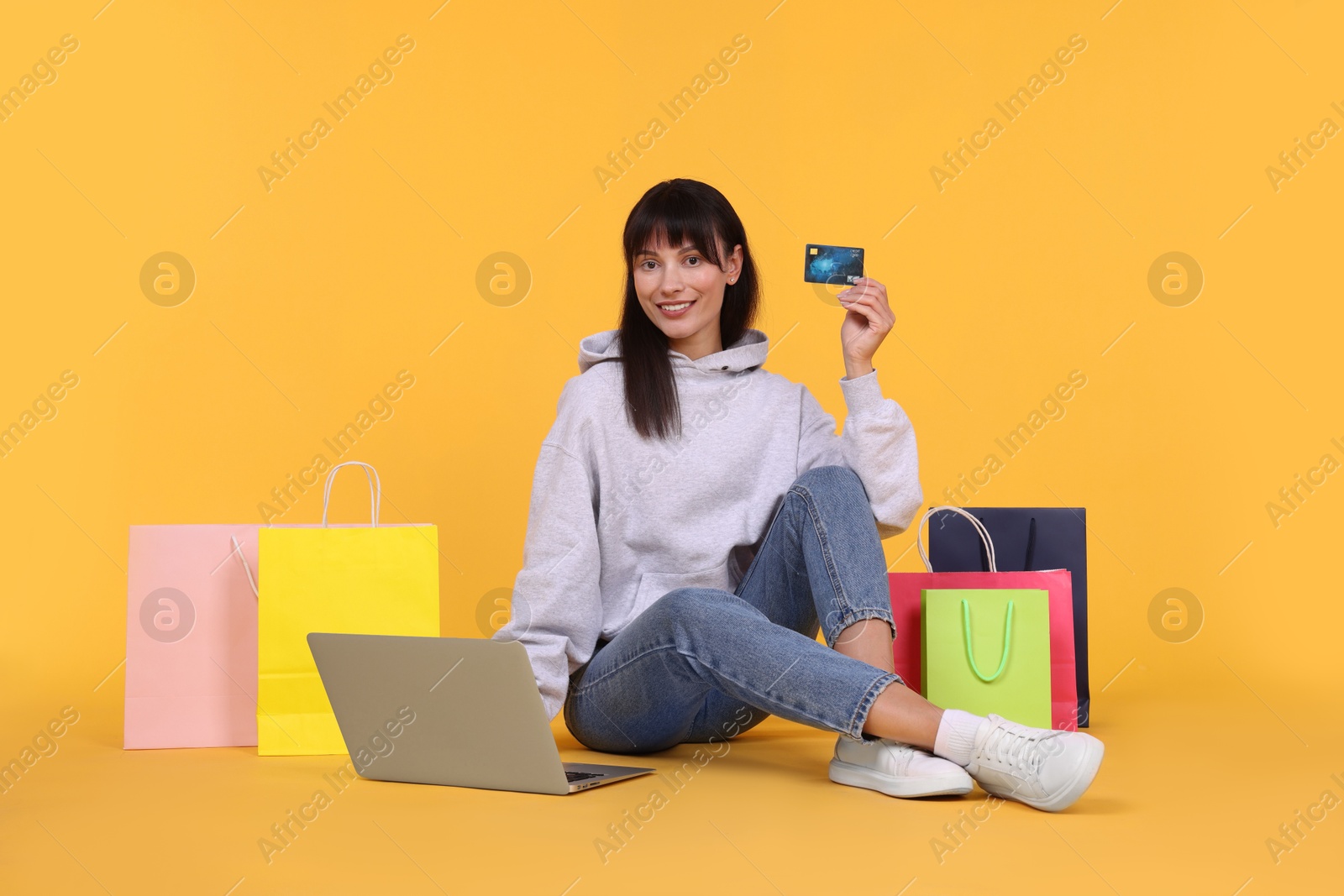 Photo of Internet shopping. Happy woman with credit card, laptop and colorful bags on orange background