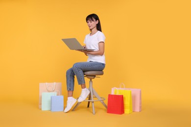 Photo of Internet shopping. Happy woman with laptop sitting on stool among colorful bags against orange background