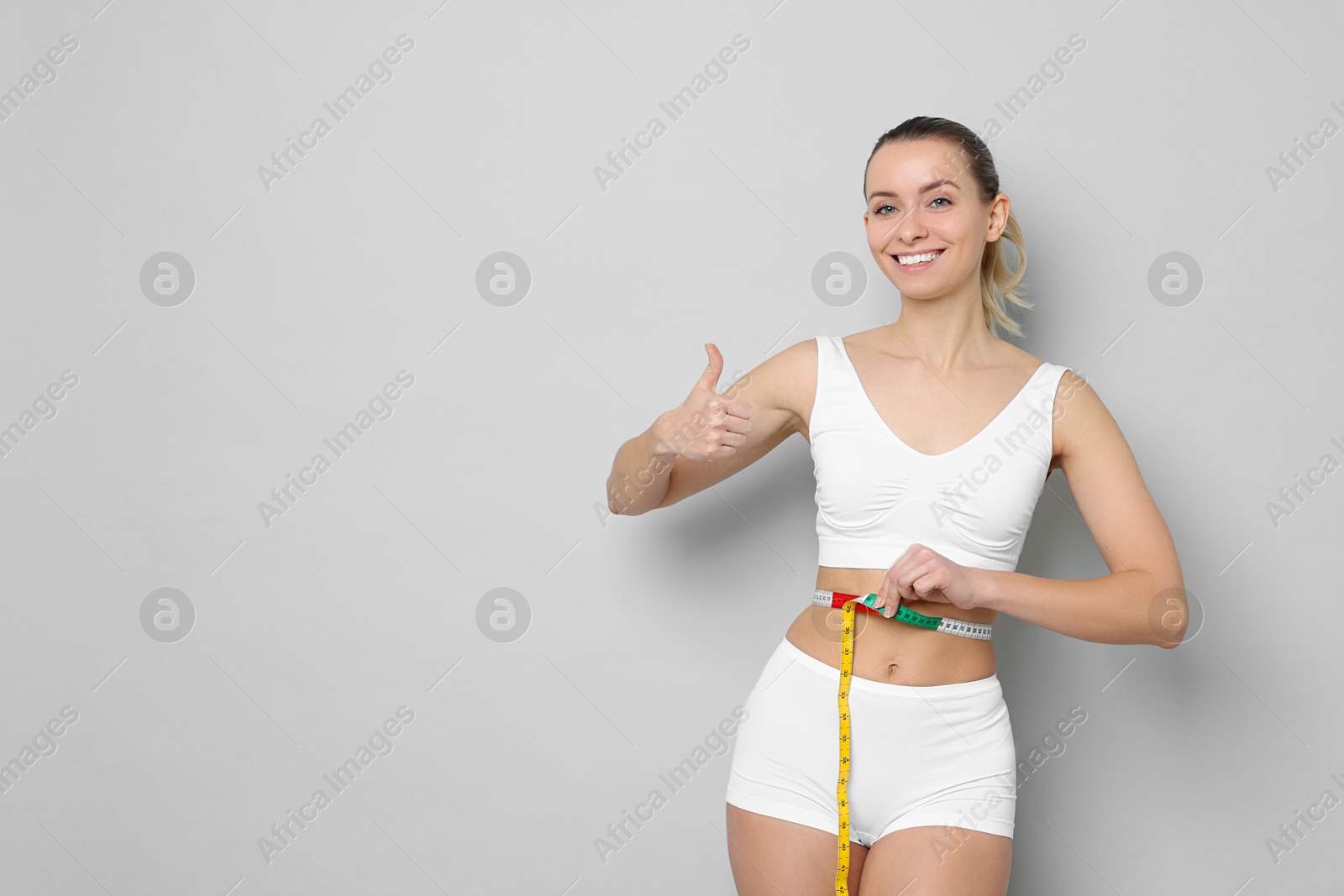 Photo of Weight loss. Happy woman measuring her slim waist with tape and showing thumbs up on light grey background, space for text