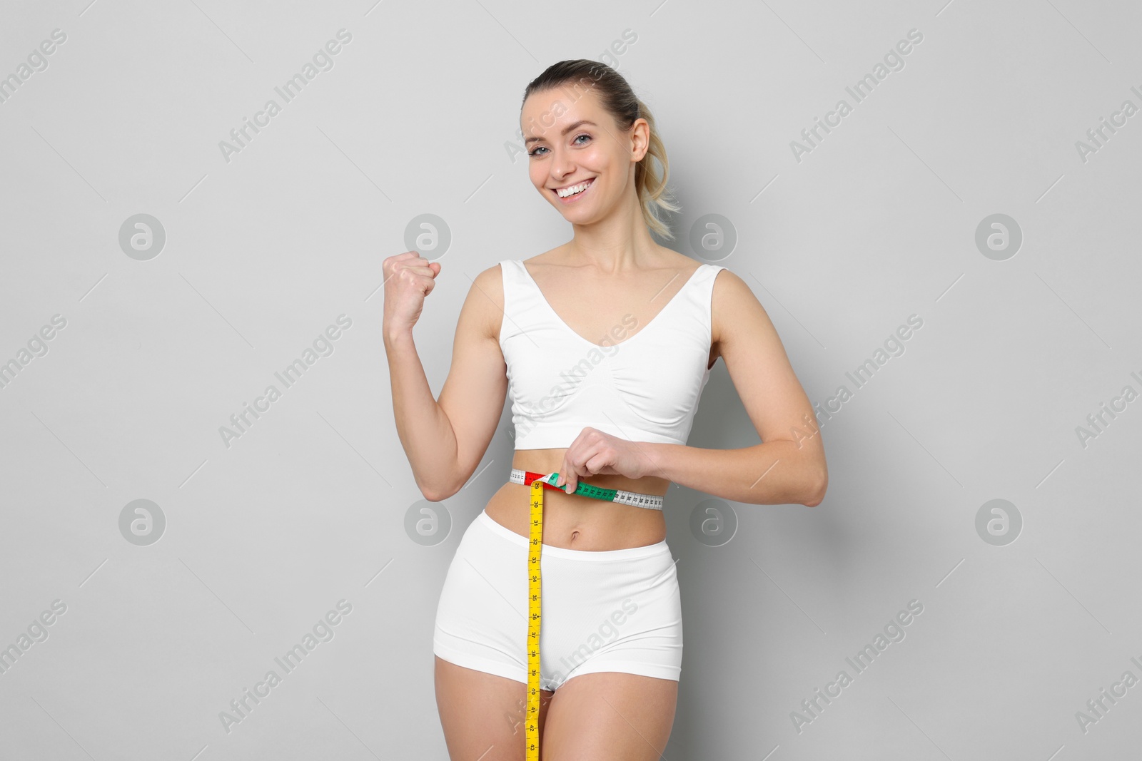 Photo of Weight loss. Happy woman measuring her slim waist with tape on light grey background