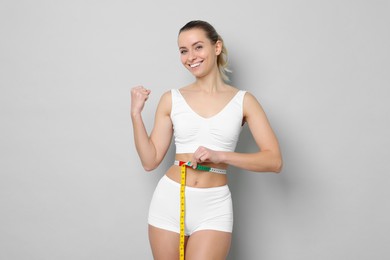 Photo of Weight loss. Happy woman measuring her slim waist with tape on light grey background