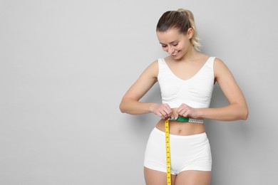 Photo of Weight loss. Happy woman measuring her slim waist with tape on light grey background, space for text