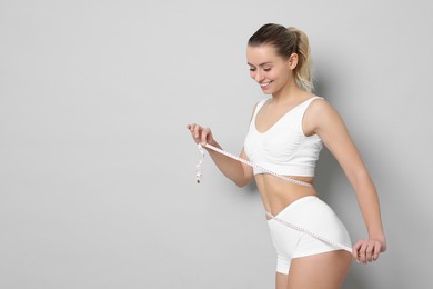 Photo of Weight loss. Happy woman measuring her slim waist with tape on light grey background, space for text