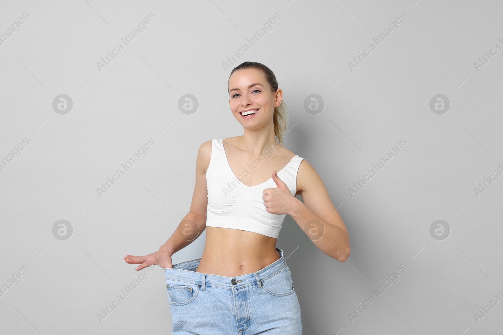 Photo of Weight loss. Happy woman wearing big jeans and showing thumbs up on light grey background