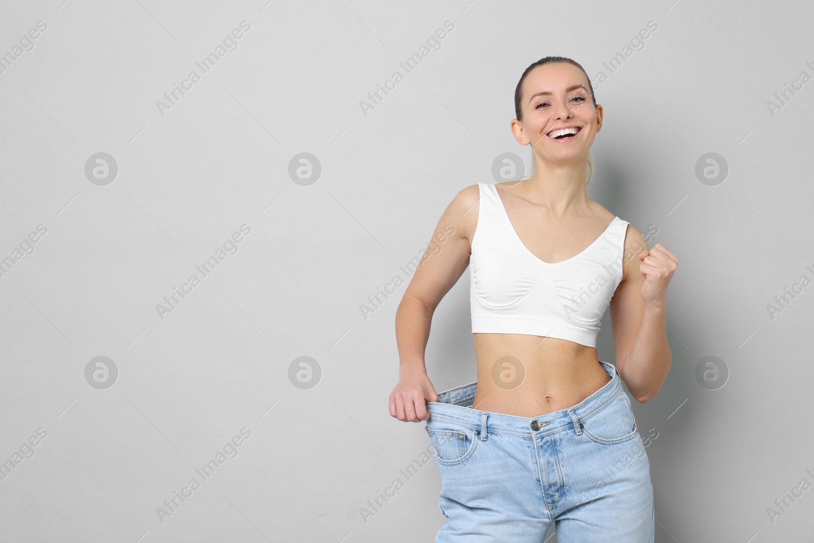 Photo of Weight loss. Happy woman wearing big jeans on light grey background, space for text