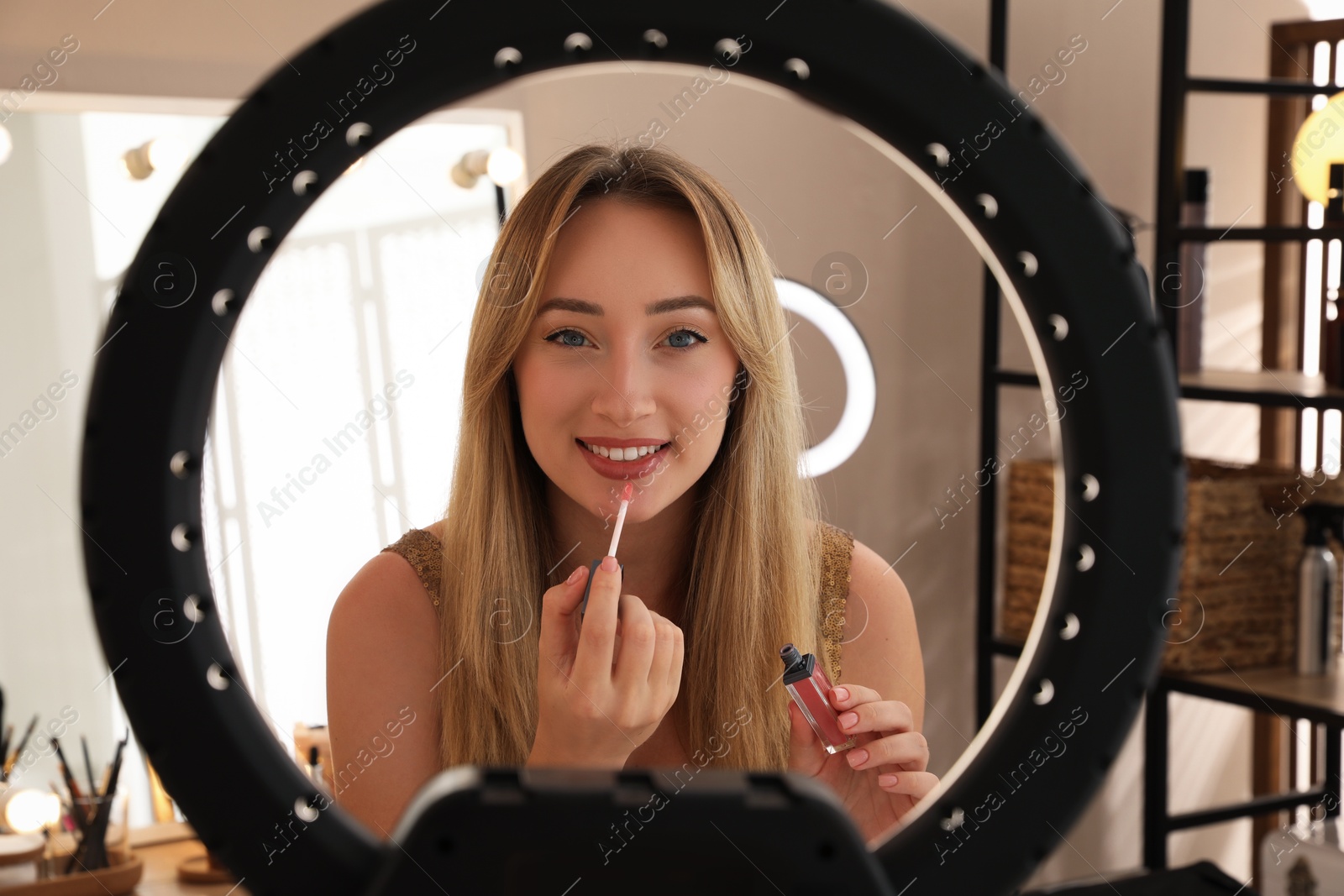 Photo of Beautiful young woman applying liquid lipstick indoors, view through ring lamp