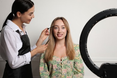 Photo of Professional makeup artist working with beautiful young woman near white wall indoors. Using ring lamp