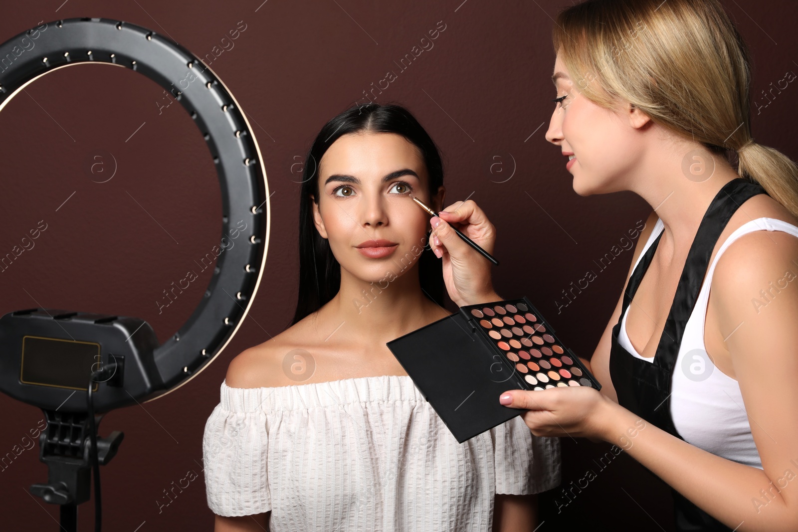 Photo of Professional makeup artist working with beautiful young woman against brown background. Using ring lamp