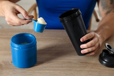 Photo of Making protein cocktail. Man with scoop of powder and shaker at wooden table, closeup