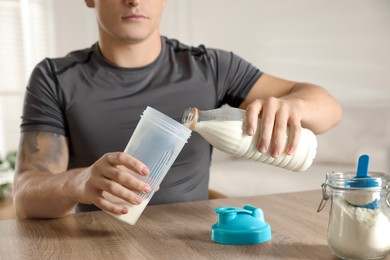 Photo of Making protein cocktail. Man pouring milk into shaker at wooden table, closeup