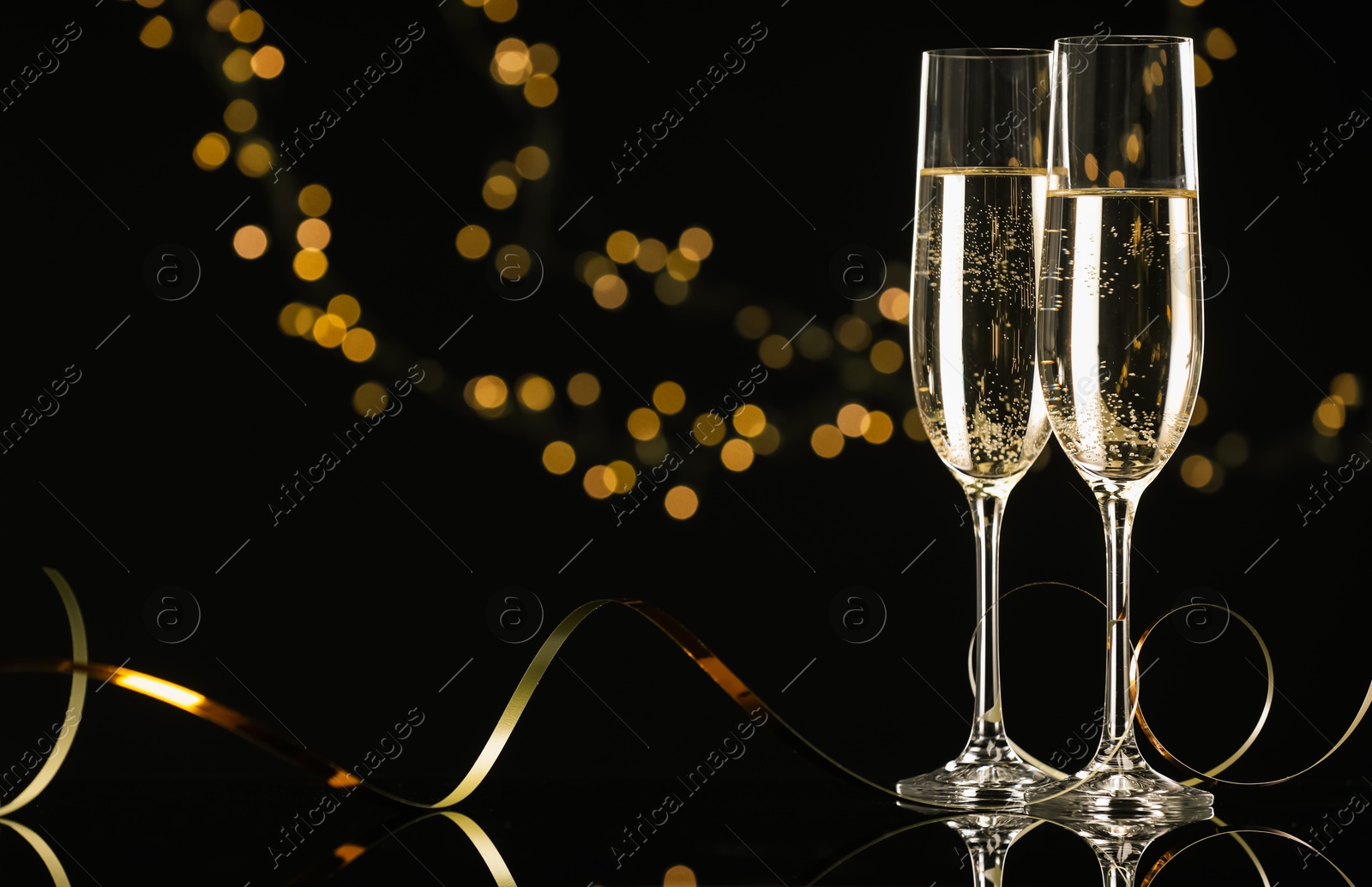 Photo of Champagne in glasses and party streamers on mirror surface against black background with blurred lights, space for text