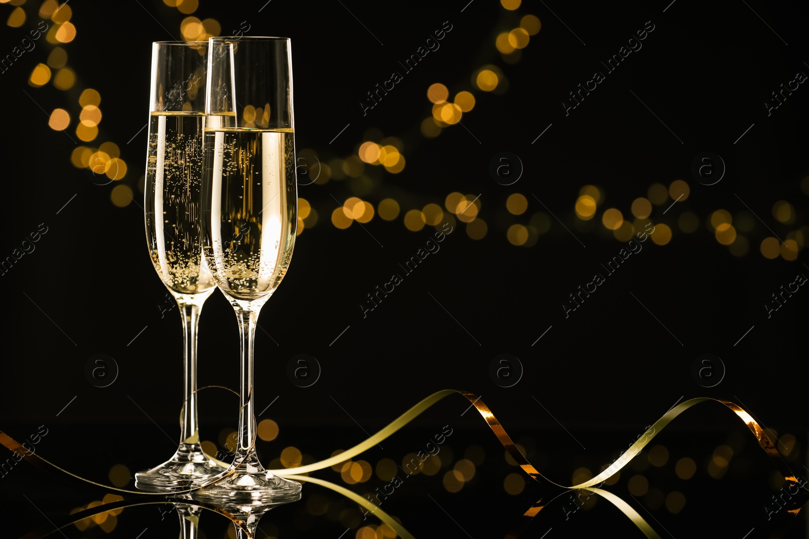 Photo of Champagne in glasses and party streamers on mirror surface against black background with blurred lights, space for text