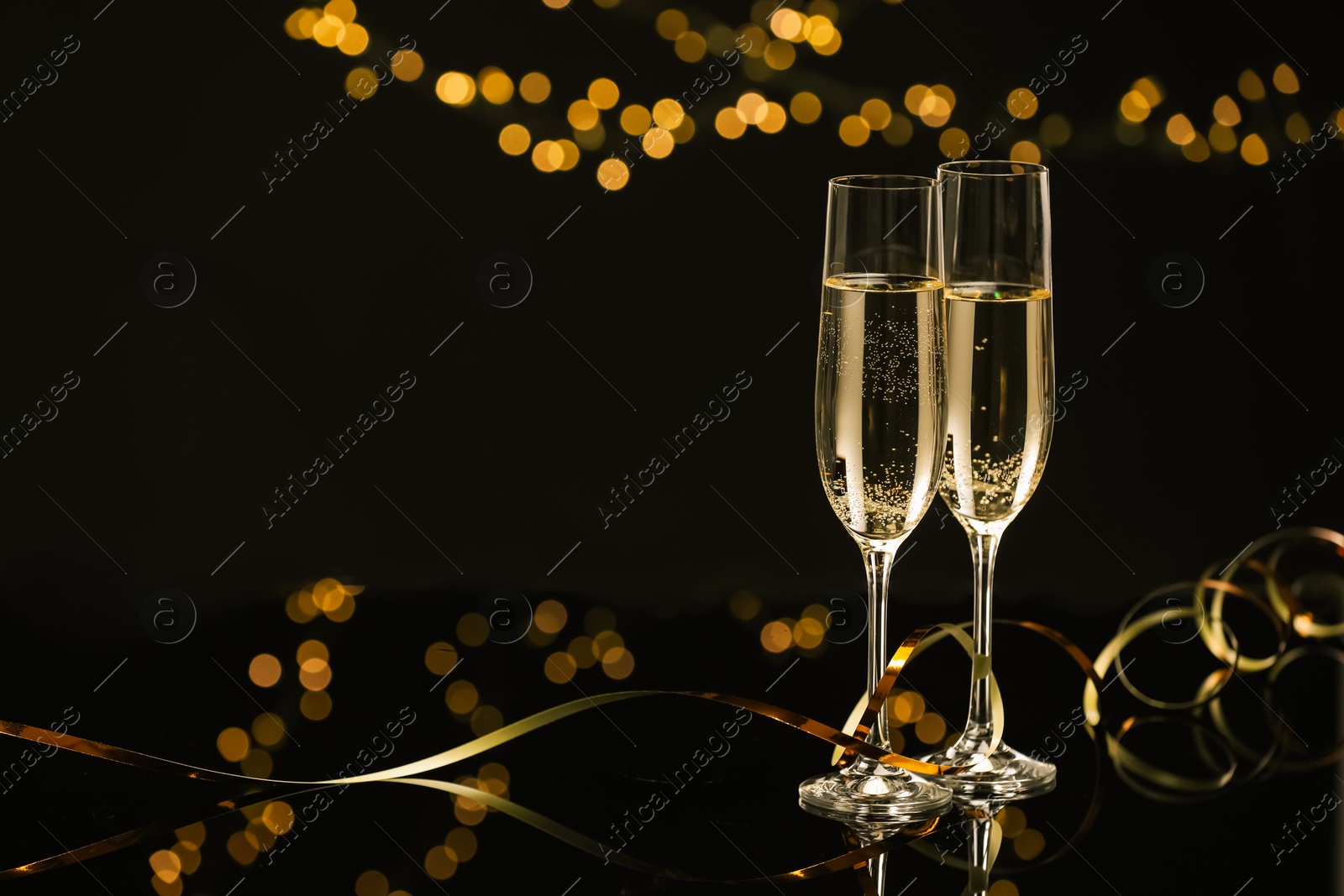 Photo of Champagne in glasses and party streamers on mirror surface against black background with blurred lights, space for text