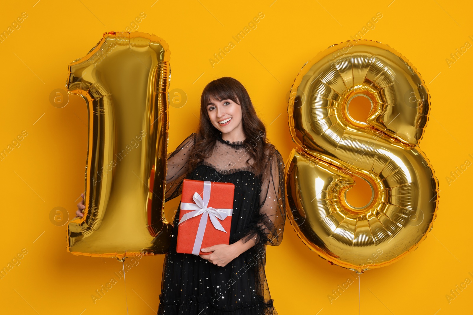 Photo of Coming of age party - 18th birthday. Young woman with number shaped balloons and gift box on yellow background