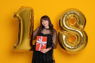 Photo of Coming of age party - 18th birthday. Young woman with number shaped balloons and gift box on yellow background