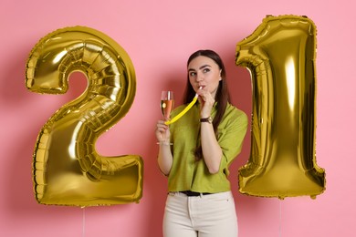 Photo of Coming of age party - 21st birthday. Happy young woman with number shaped balloons, glass of sparkling wine and blower on pink background