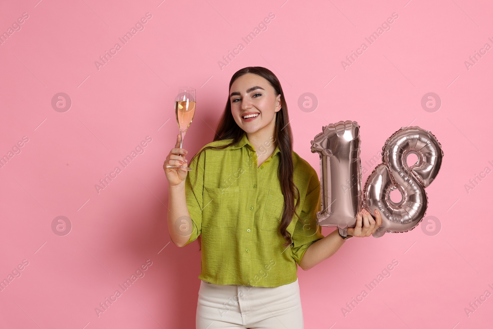 Photo of Coming of age party - 18th birthday. Happy young woman with number shaped balloons and glass of sparkling wine on pink background