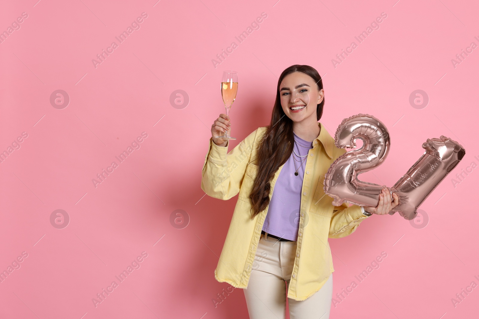 Photo of Coming of age party - 21st birthday. Happy young woman with number shaped balloons and glass of sparkling wine on pink background, space for text