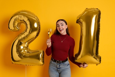 Photo of Coming of age party - 21st birthday. Happy young woman with number shaped balloons and glass of sparkling wine on yellow background