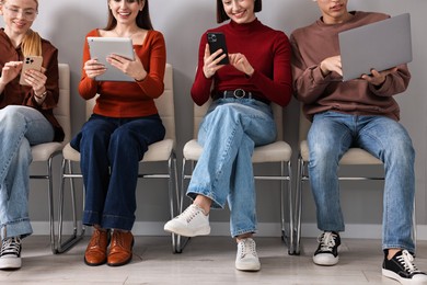 Photo of Group of people using different gadgets on chairs near grey wall indoors. Modern technology