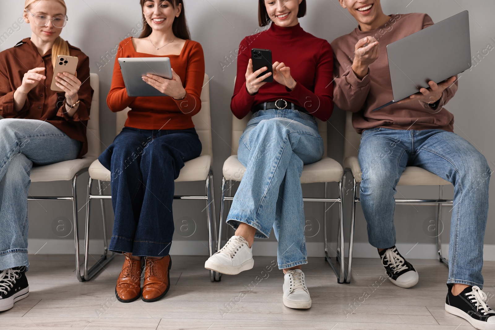 Photo of Group of people using different gadgets on chairs near grey wall indoors. Modern technology