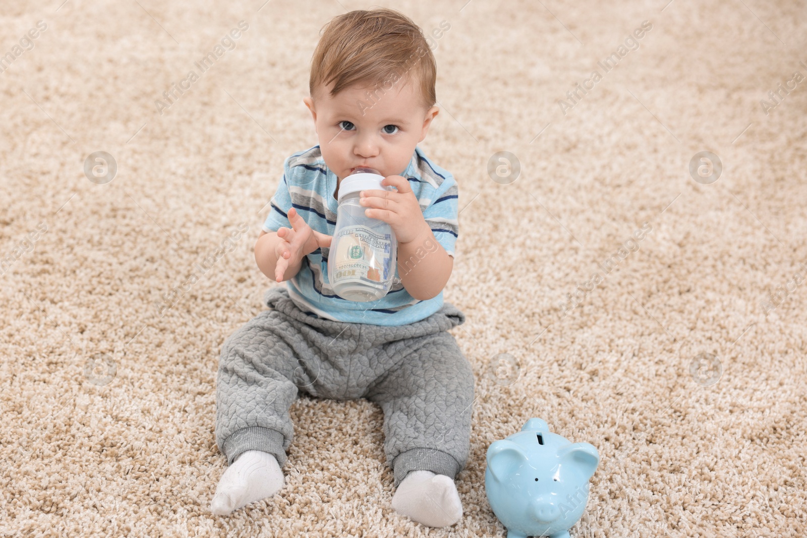 Photo of Cute little baby with money in bottle and piggybank on floor at home