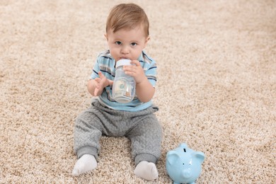 Photo of Cute little baby with money in bottle and piggybank on floor at home