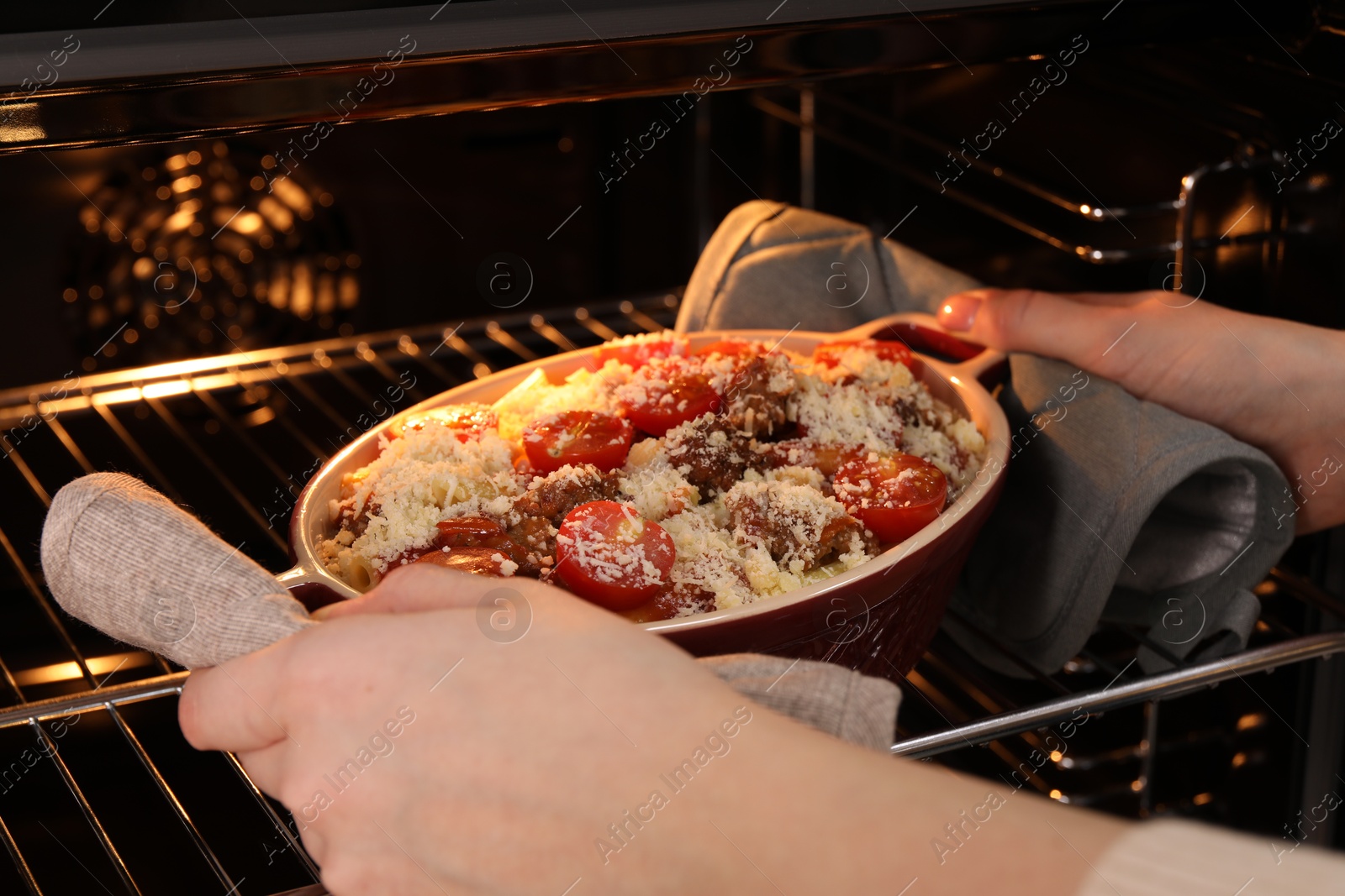 Photo of Woman putting pasta casserole with cheese, tomatoes and minced meat into oven, closeup