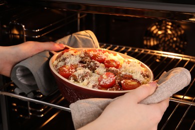 Photo of Woman putting pasta casserole with cheese, tomatoes and minced meat into oven, closeup