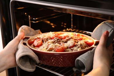 Photo of Woman putting pasta casserole with cheese, tomatoes and minced meat into oven, closeup