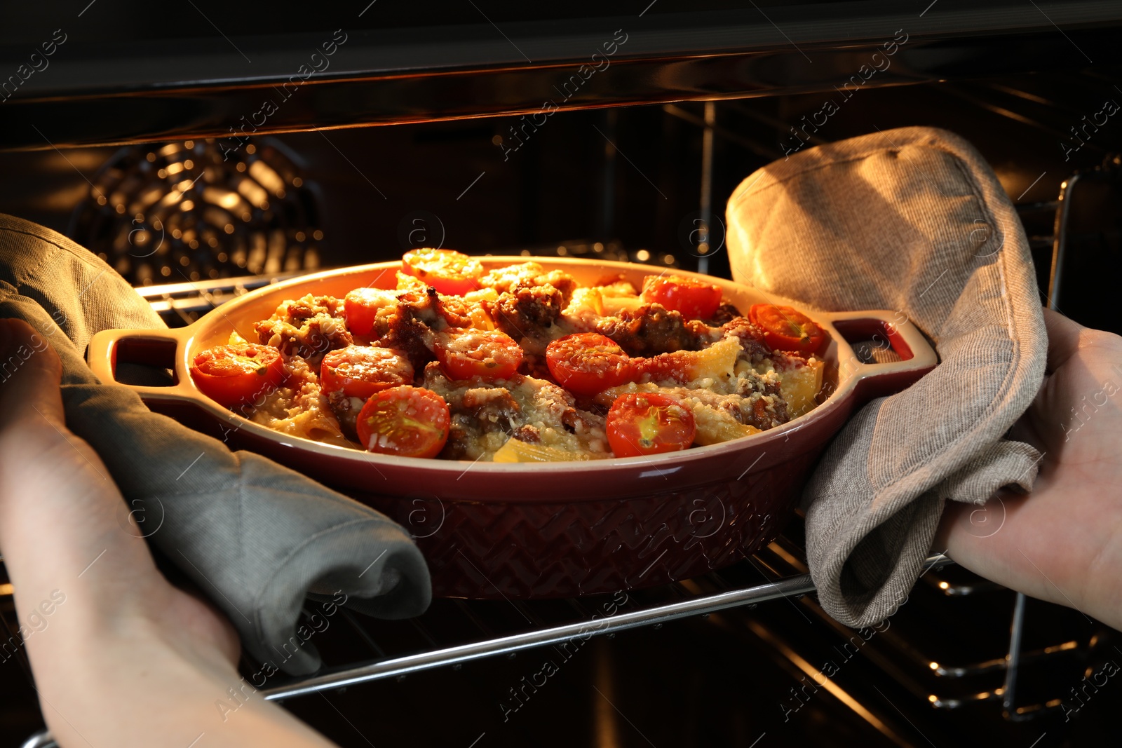 Photo of Woman taking delicious pasta casserole with cheese, tomatoes and minced meat out of oven, closeup