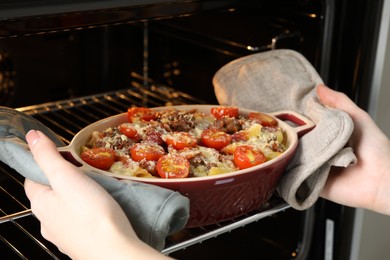 Photo of Woman taking delicious pasta casserole with cheese, tomatoes and minced meat out of oven, closeup