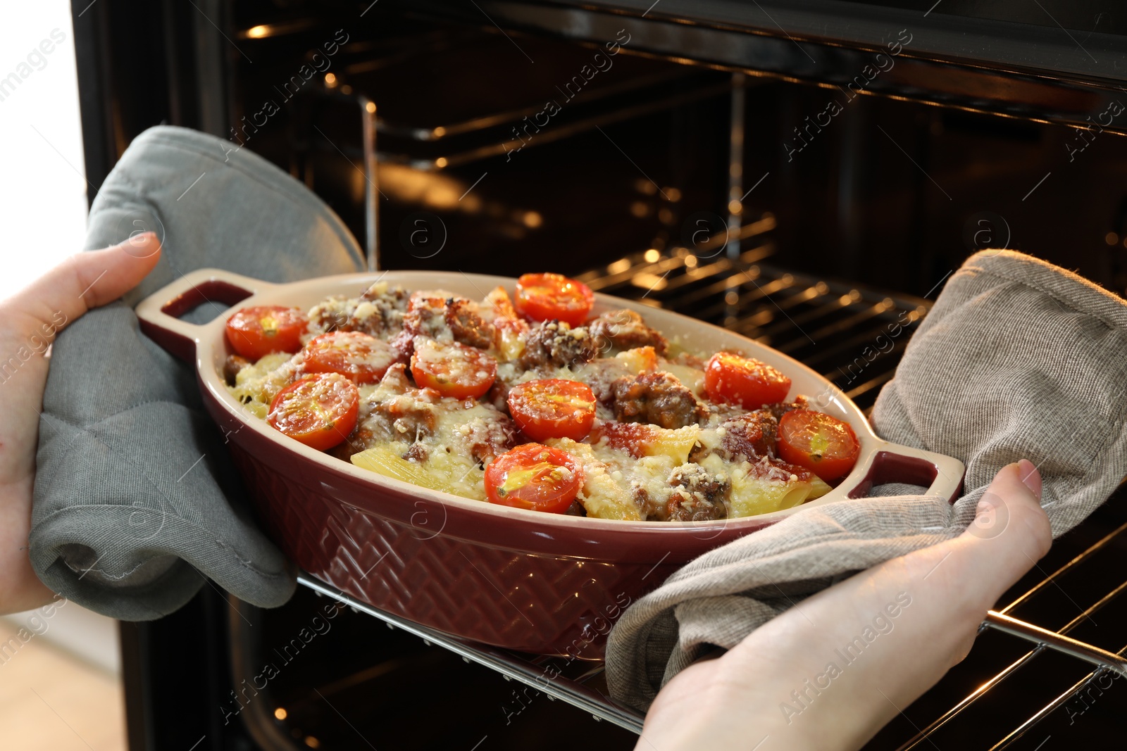 Photo of Woman taking delicious pasta casserole with cheese, tomatoes and minced meat out of oven, closeup