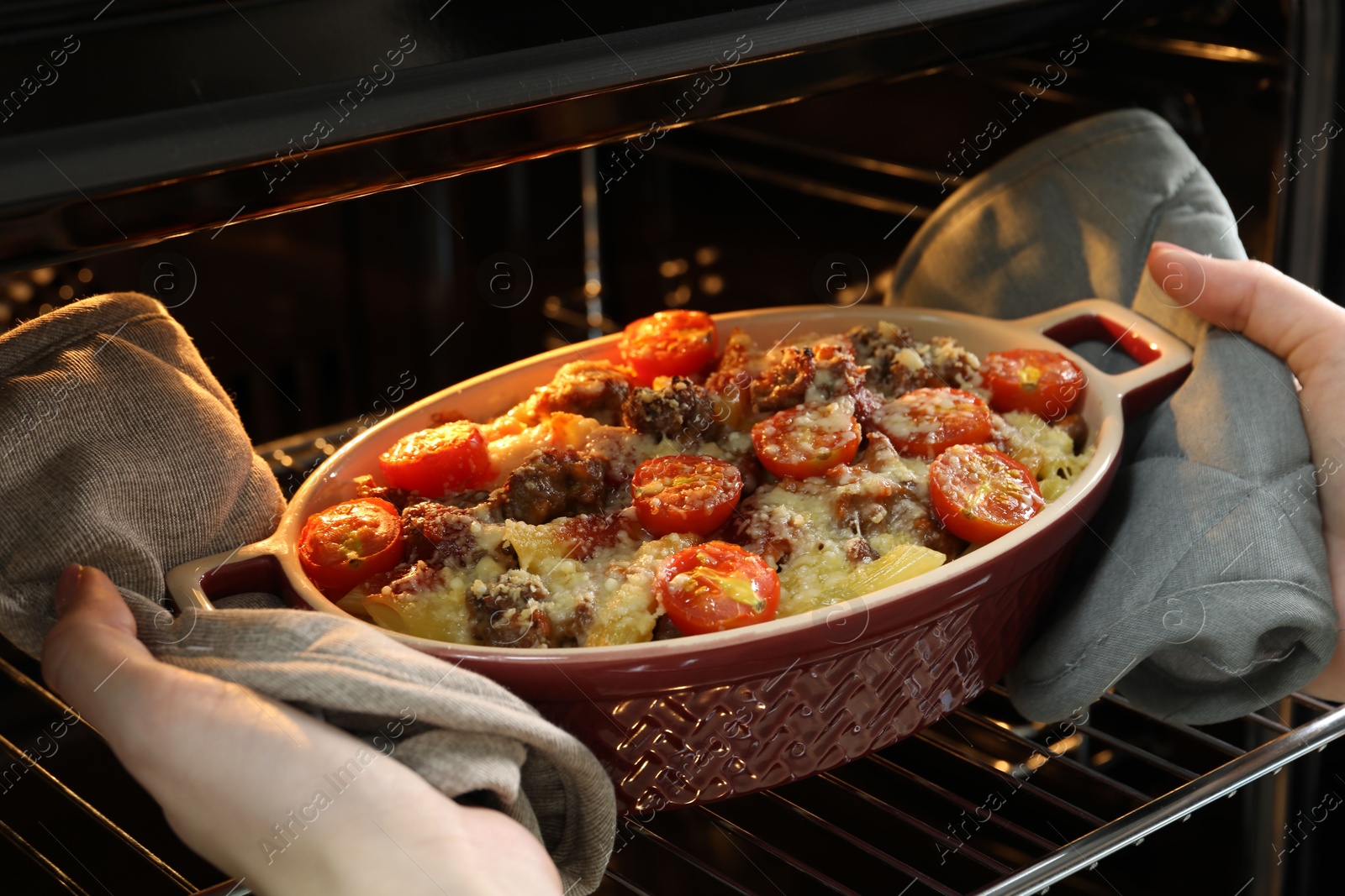 Photo of Woman taking delicious pasta casserole with cheese, tomatoes and minced meat out of oven, closeup