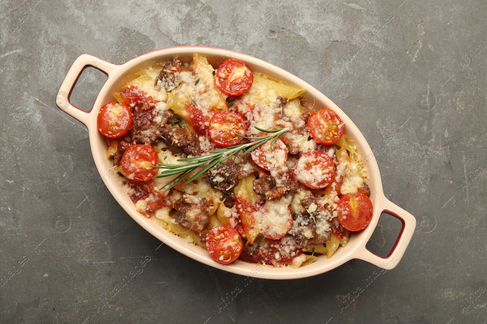 Photo of Delicious pasta casserole with cheese, tomatoes, minced meat and rosemary in baking dish on grey textured table, top view