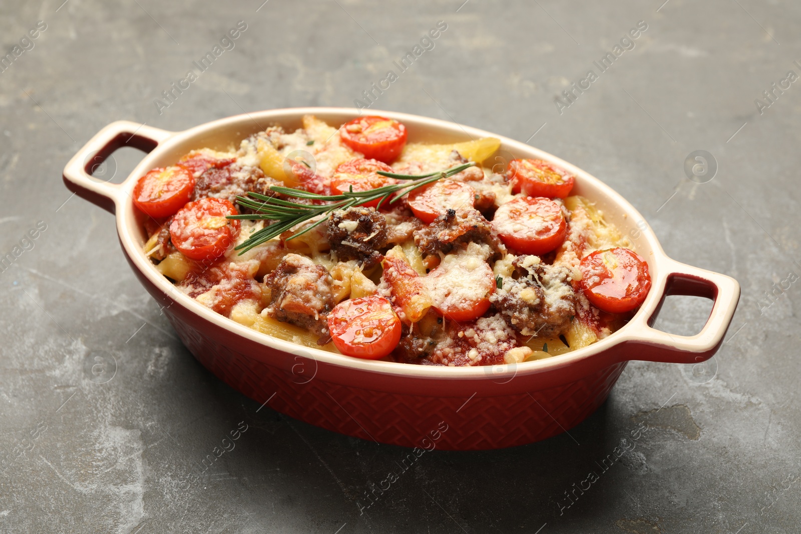 Photo of Delicious pasta casserole with cheese, tomatoes, minced meat and rosemary in baking dish on grey textured table, closeup