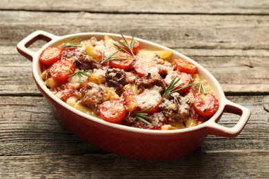 Photo of Delicious pasta casserole with cheese, tomatoes, minced meat and rosemary in baking dish on wooden table, closeup