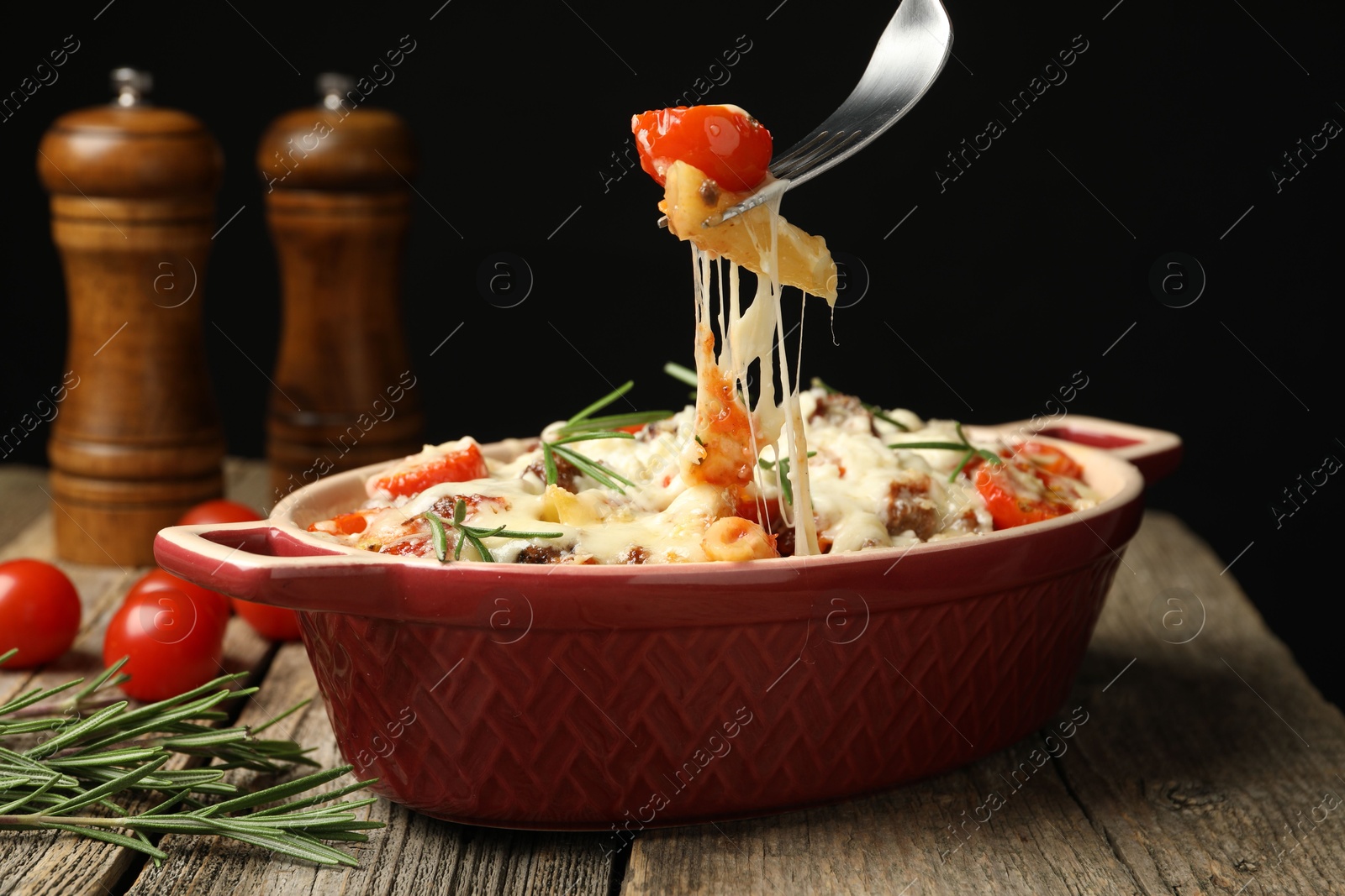 Photo of Eating delicious pasta casserole with fork at wooden table against black background, closeup