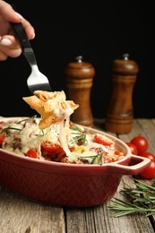 Photo of Woman eating delicious pasta casserole with fork at wooden table against black background, closeup