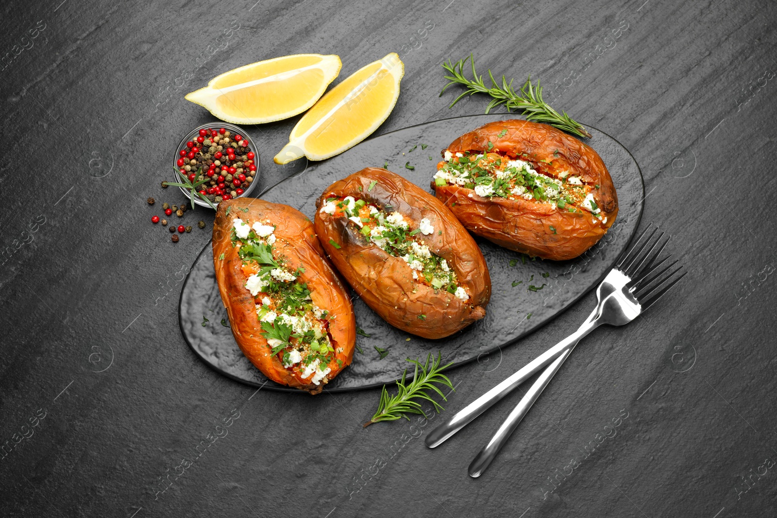 Photo of Tasty baked sweet potato with feta cheese, lemon, peppercorns and herbs on dark textured table, flat lay