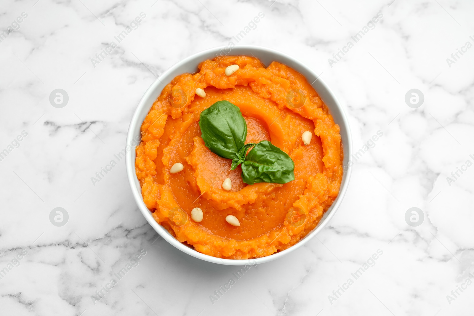 Photo of Tasty mashed sweet potato with basil and seeds in bowl on white marble table, top view