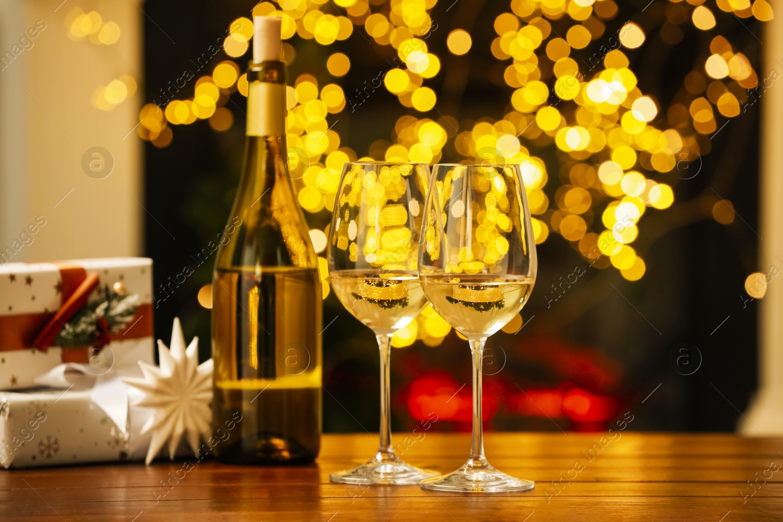 Photo of Glasses and bottle of white wine on wooden table against blurred Christmas lights. Bokeh effect