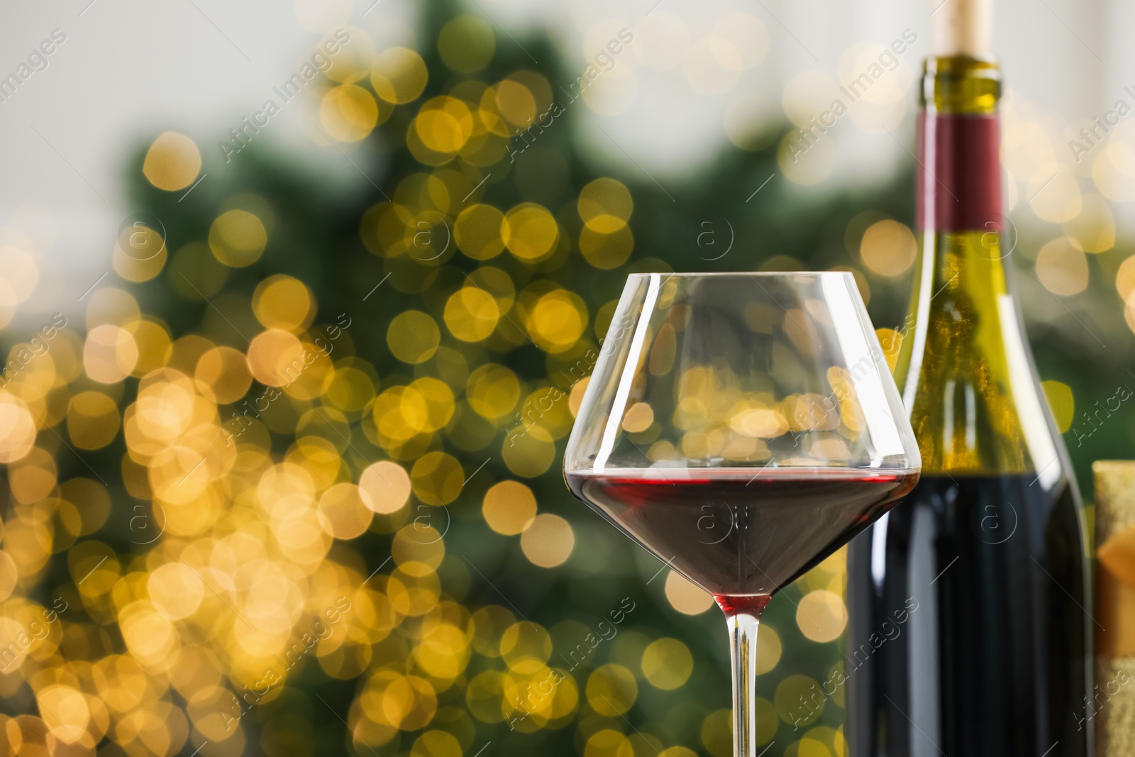 Photo of Glass and bottle of red wine against blurred Christmas lights, closeup. Bokeh effect